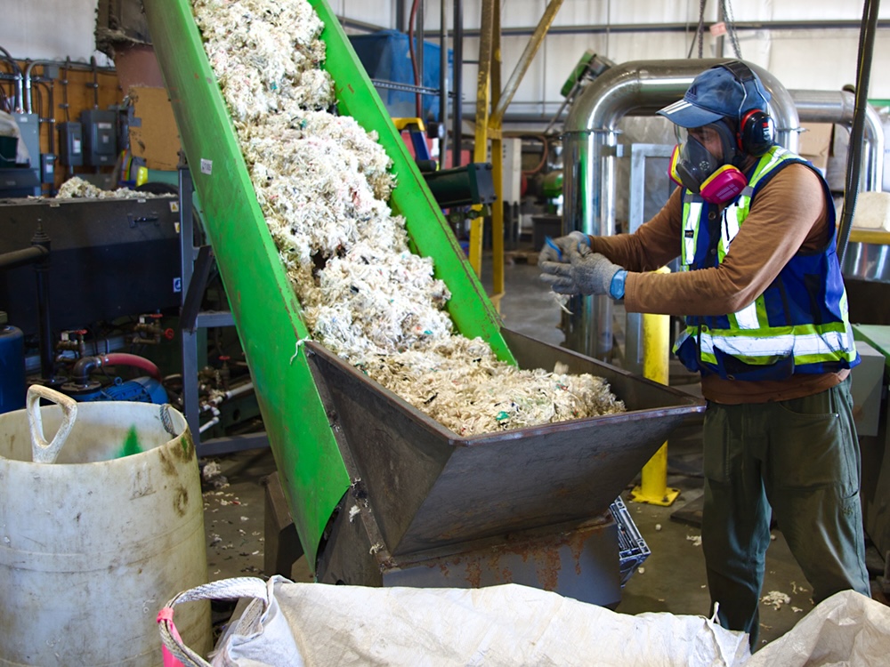 A person wearing a clear face mask, pink respirator, black over-ear protection, blue baseball hat and blue and yellow reflective safety vest feeds shredded nylon onto a conveyer belt.