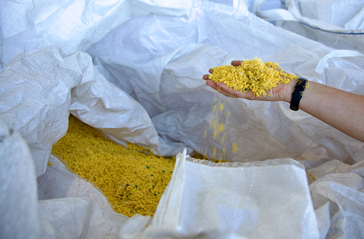 A light-skin-tone forearm extends into the frame, the hand cupped to hold shredded yellow nylon rope. Below the arm is an extra large white plastic bag full of shredded yellow rope.