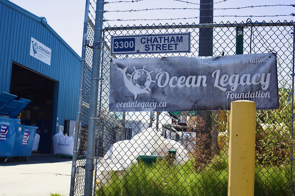 A faded black and white banner on a chain-link fence reads ‘The Ocean Legacy Foundation, Oceanlegacy.ca.’ Beyond the fence is a two-storey blue corrugated metal warehouse.