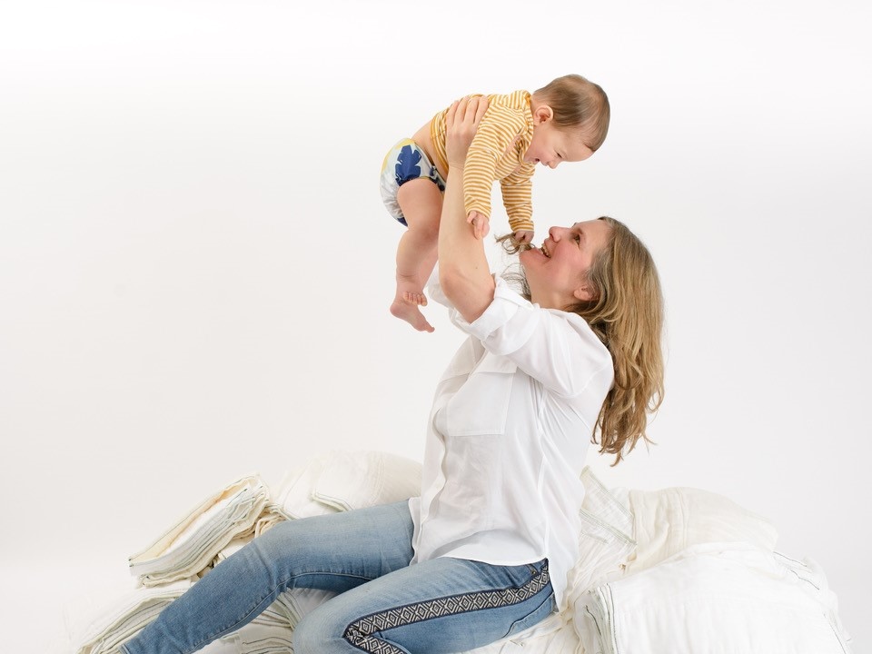 A white woman with blond shoulder-length hair wearing a white blouse smiles at the brown-haired baby she holds over her head.