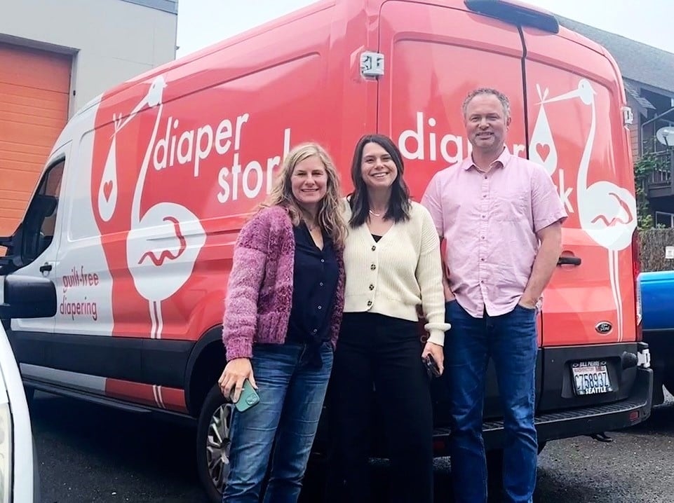 A blond white woman, a dark-haired white woman (both in their late 30s) and a white man with salt-and-pepper hair stand in front of a delivery van that says ‘diaper stork’ on its side. 