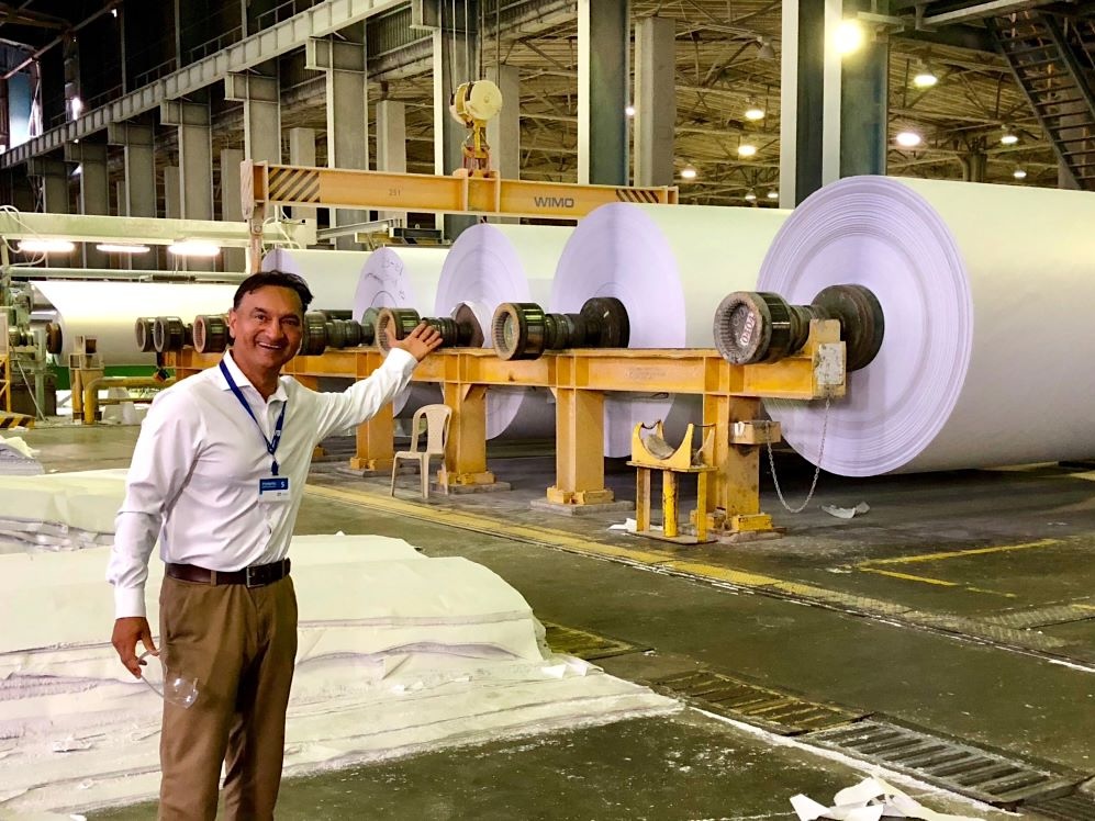 A man with medium skin tone and short dark hair stands in a paper factory, gesturing towards very large rolls of paper.