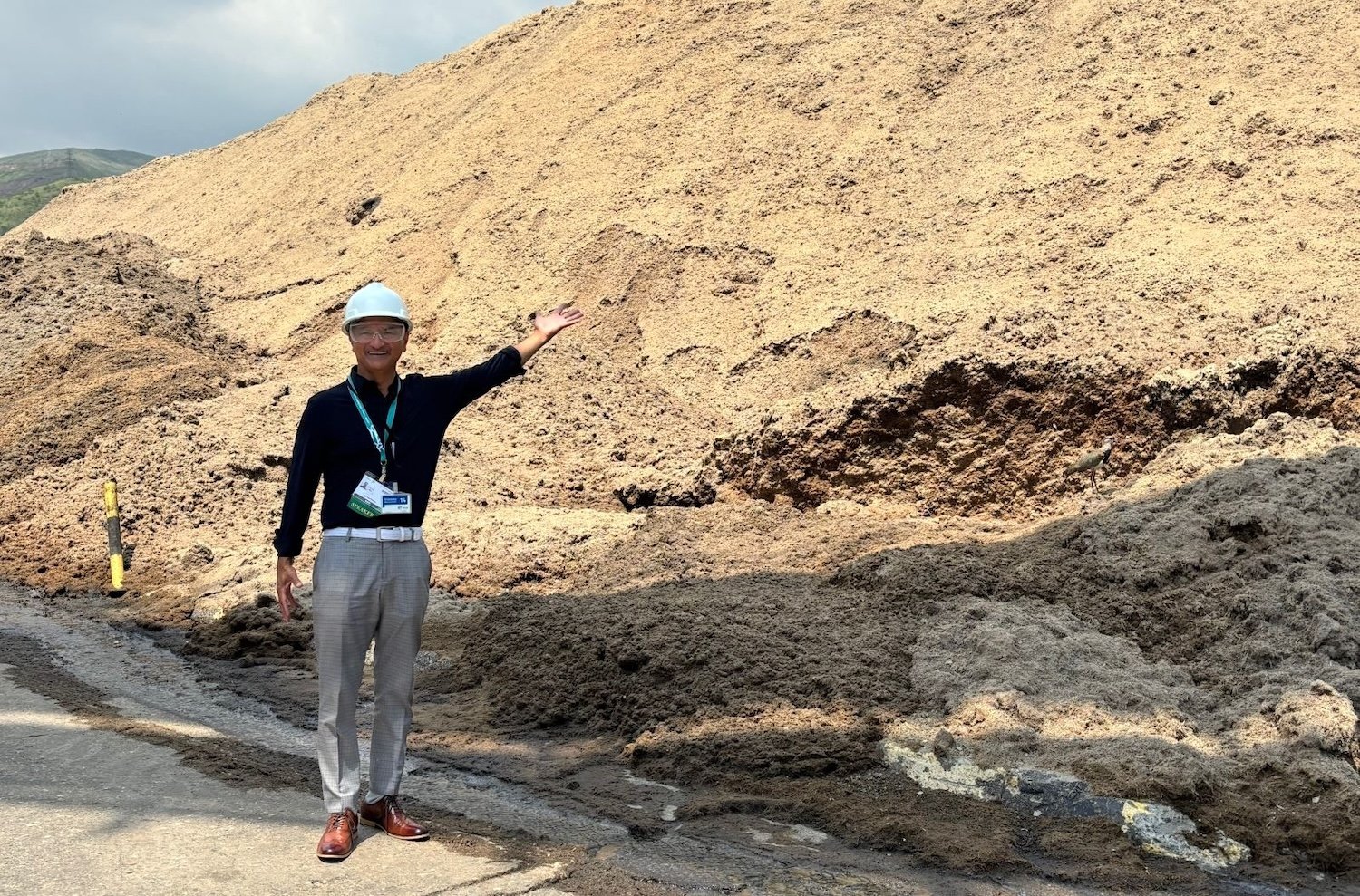 A man wearing a hard hat stands in front of a large mound of brownish stuff that looks sort of like sawdust. He gestures towards it.