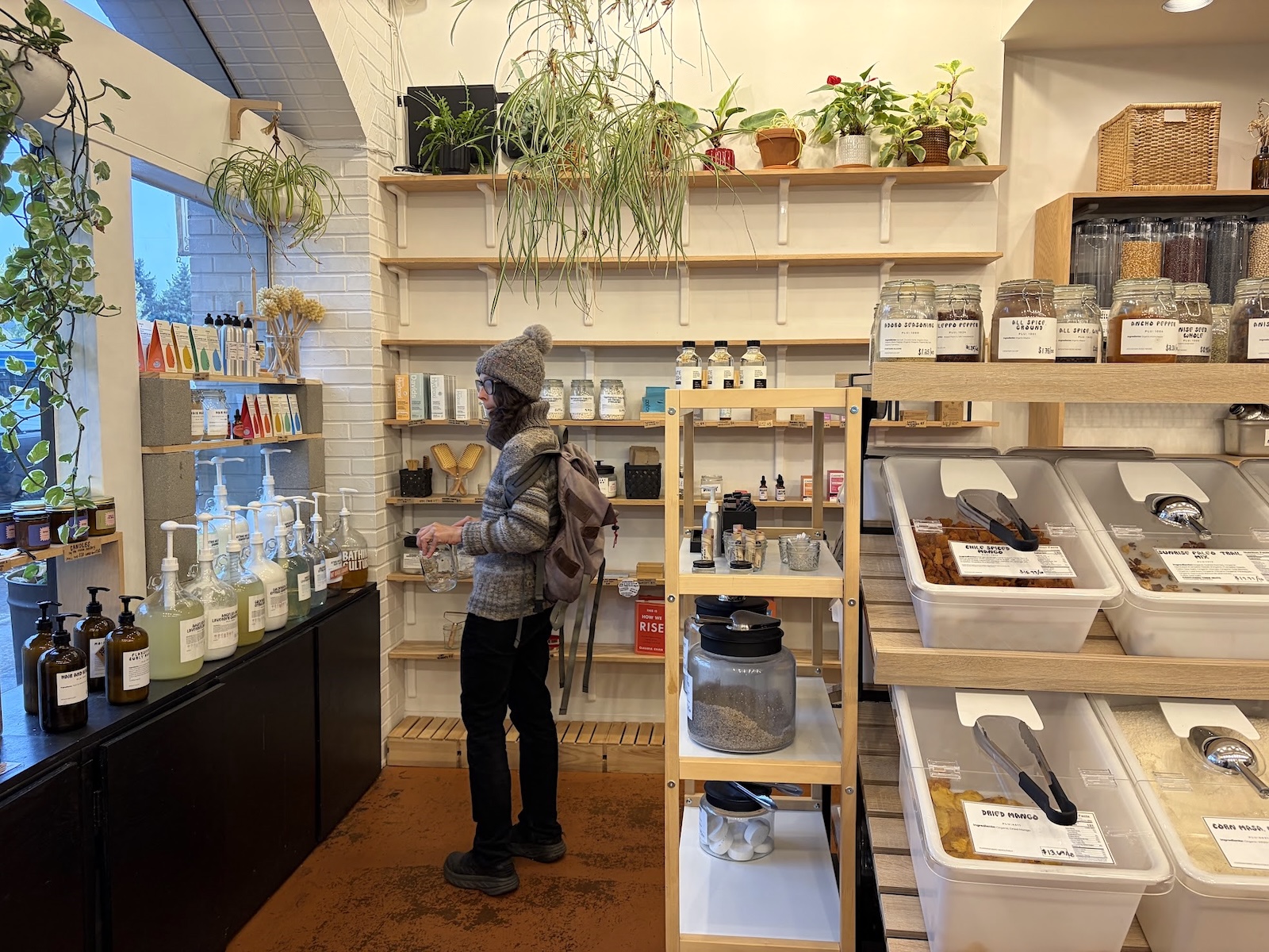 A person glances at the shelves of a refillery. There are plants and multiple boxes of goods for purchase. 