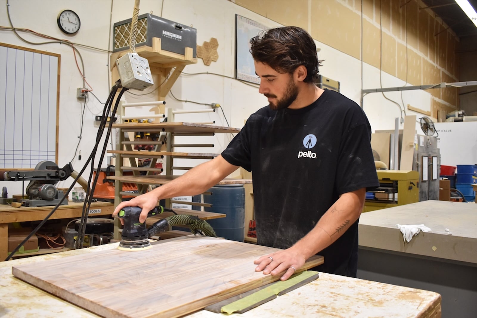 A man in a black T-shirt in a wood shop uses a sanding machine.