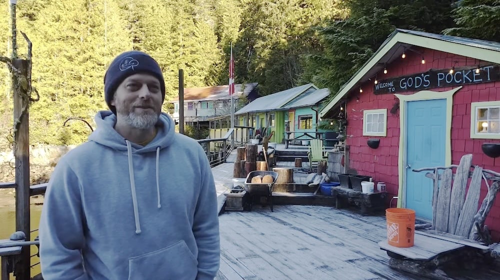 A man in a toque stands on a dock. A building with the words ‘God’s Pocket’ is just over his left shoulder.