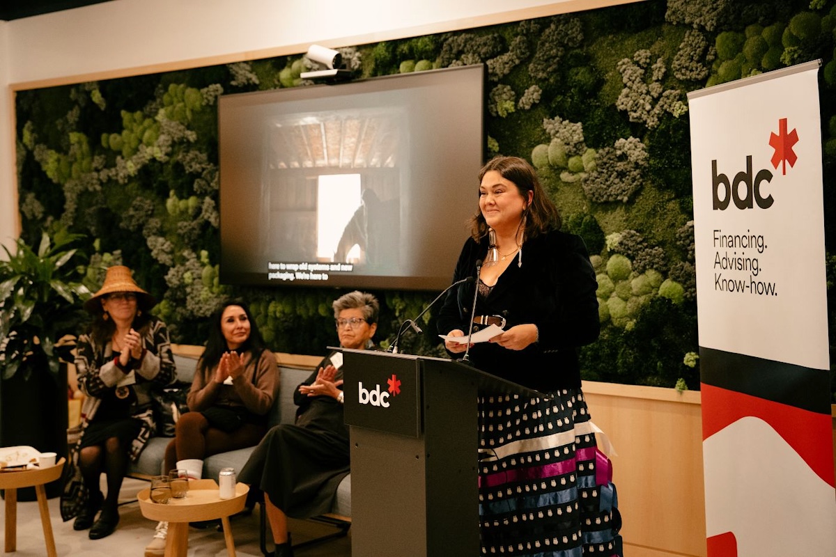 A woman stands in front of a podium in a ballroom. A BDC board and screen is in the background.