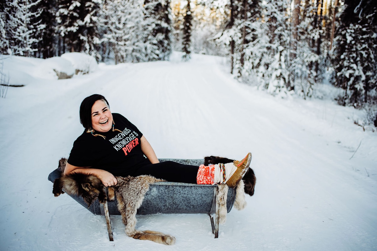 A woman sits in the snow, wearing a T-shirt that says, ‘Indigenous Knowledge is Power.’