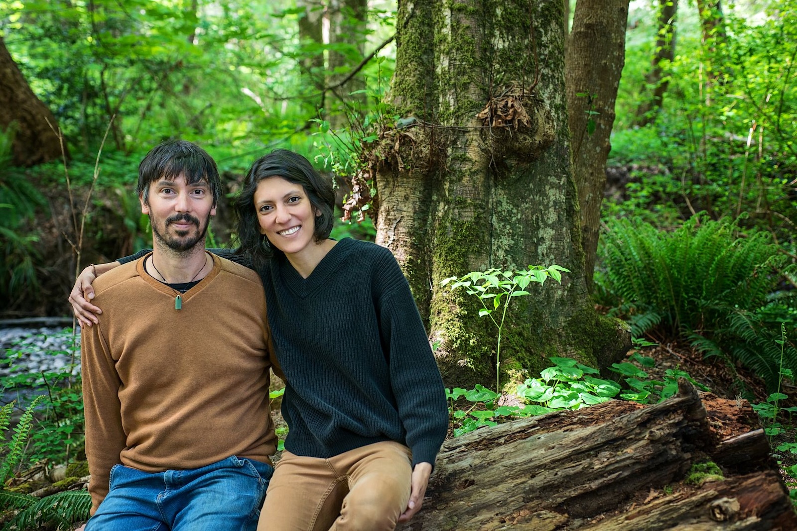 A bearded young man and a smiling young woman with black hair sit on a log in a verdant rain forest. 