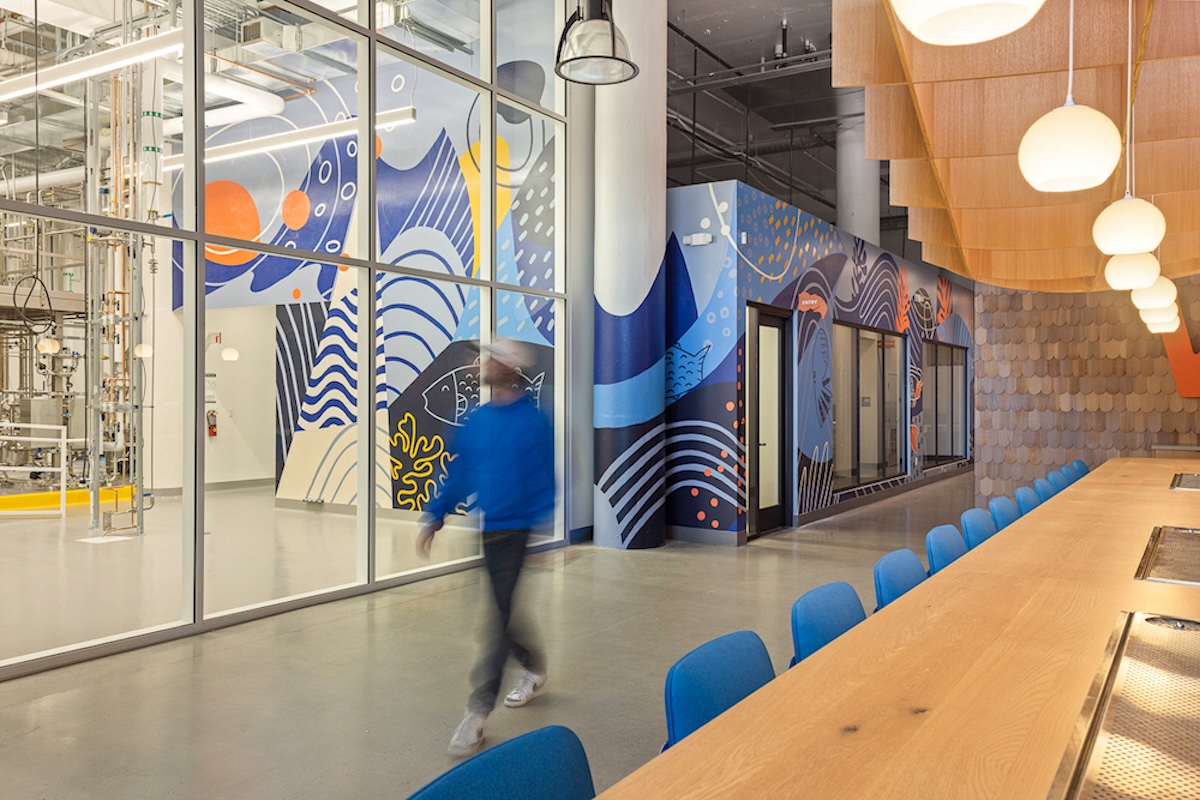 A man walks through a microbrewery facility known as the Fishery. He walks beside a long wooden table. There&rsquo;s a multi-coloured wall and industrial-looking machines in the background. 