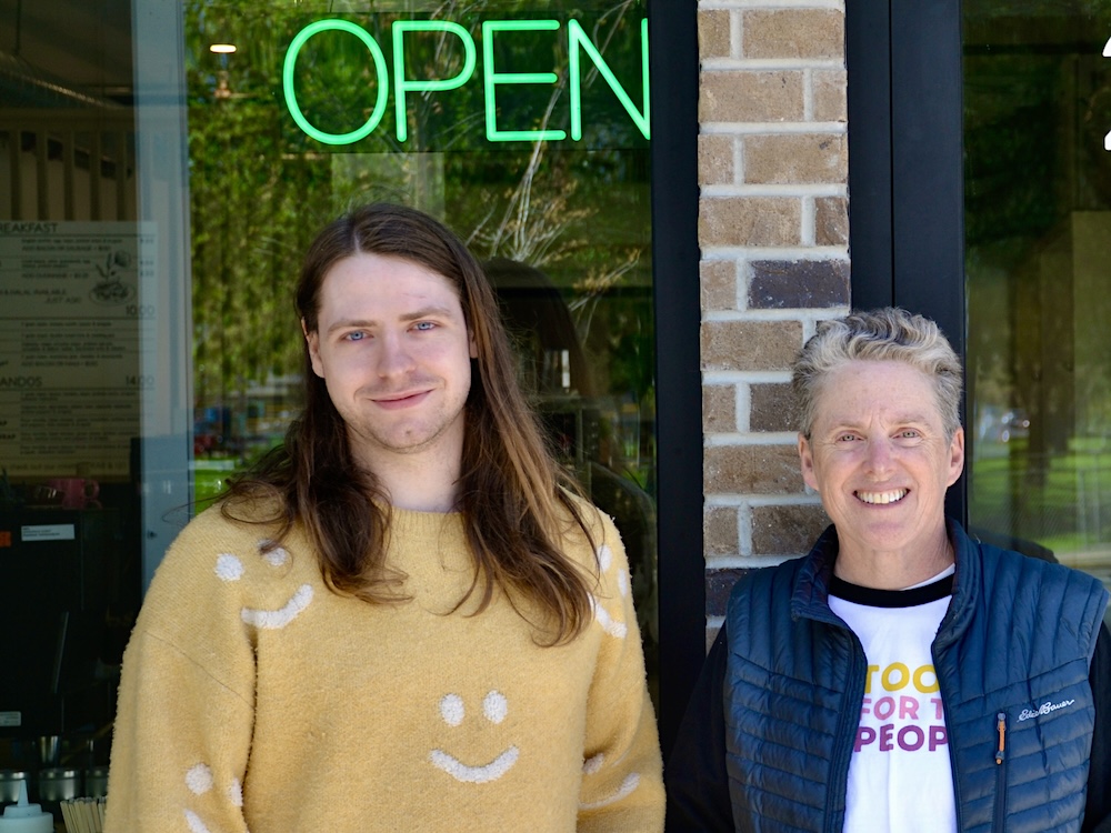 Chance McClendon, left, wears a yellow shirt with smiley faces on it. He stands beside Jenny Farkas, who is wearing a blue vest and a shirt with the words &lsquo;Tools for the People&rsquo; on it. They are in front of a door and brick wall with an &lsquo;Open&rsquo; sign.