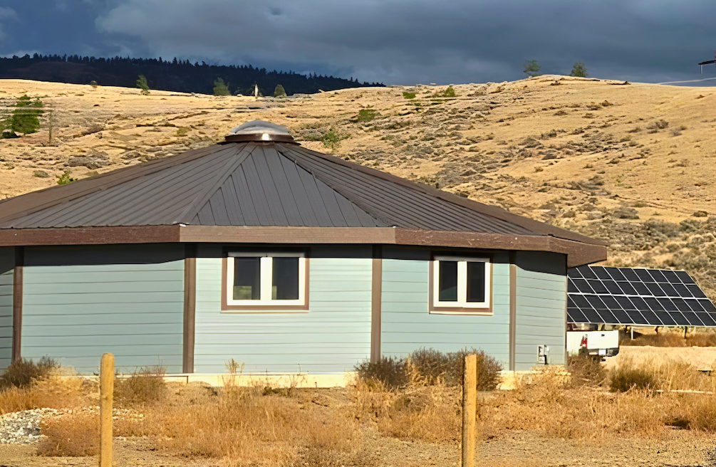 A Dodeca-Home sits on a hillside in the interior of B.C. The walls are blue and the roof is brow. There&rsquo;s a solar panel to the right of the house and brown grass and shrubs surrounding the home. 