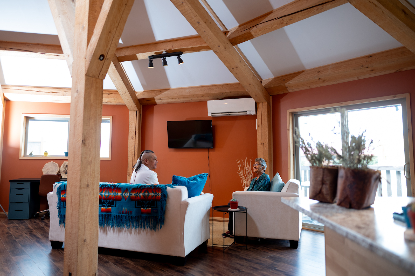 A mother and son have a chat inside a Dodeca home. The son, Robert Simon, is wearing a white t-shirt while his mother, Christine Simon, sports a turquoise long sleeve. There&rsquo;s a blanket on the couch and a television hanging on the wall. The walls are orange. 