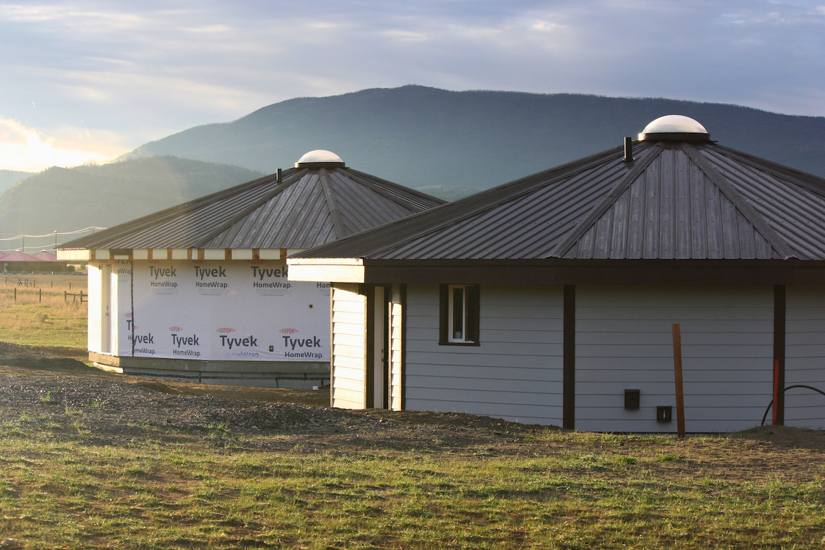 Two Dodeca-Homes stand side-by-side in a field with mountains, blue sky and sunshine in the background. The homes have a circular design, with a black-panelled roof and circular skylight. The home in the foreground has wooden panels, while the one in the distance is still in Tyvek home wrap. 