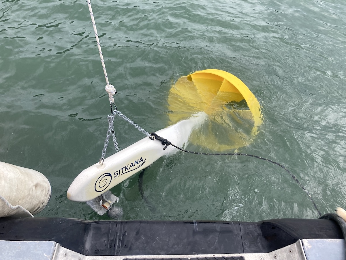 A Sitkana power-generating ocean device is placed in the water off the back of a boat. It has a bright yellow turbine and is attached to a white front piece that has the word &lsquo;Sitkana&rsquo; written on it. 